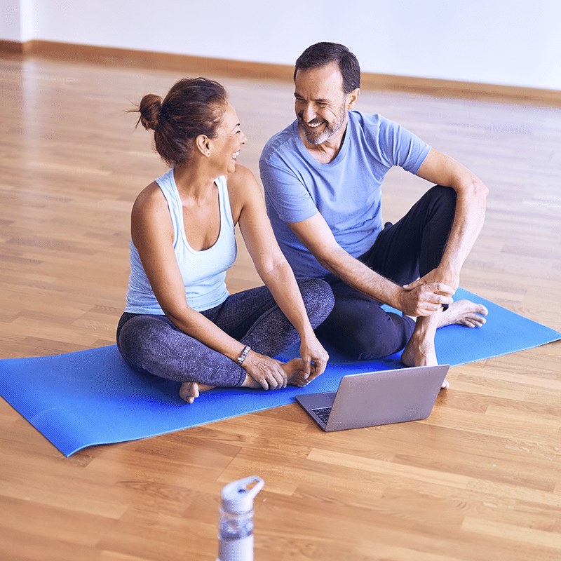 Casal sentados em tapete de yoga, com um laptop na frente deles. Eles estão virados um para o outro, sorrindo.